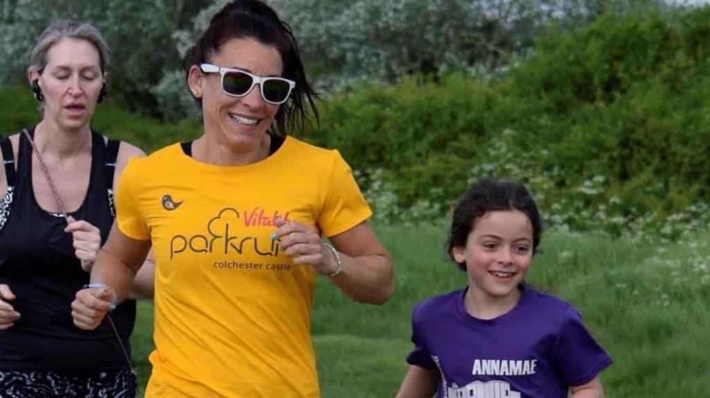 A smiling woman, wearing a yellow t-shirt, and a girl, wearing a purple t-shirt, on a run.