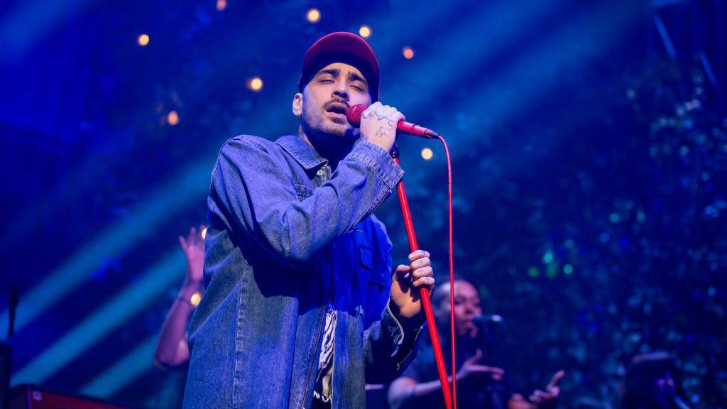 Zayn Malik performs at O2 Academy Leeds. He is wearing a blue denim shirt and a red cap. He is holding a red microphone on a red microphone stand and singing with his eyes closed. There are two background singers. The stage lighting is blue. 