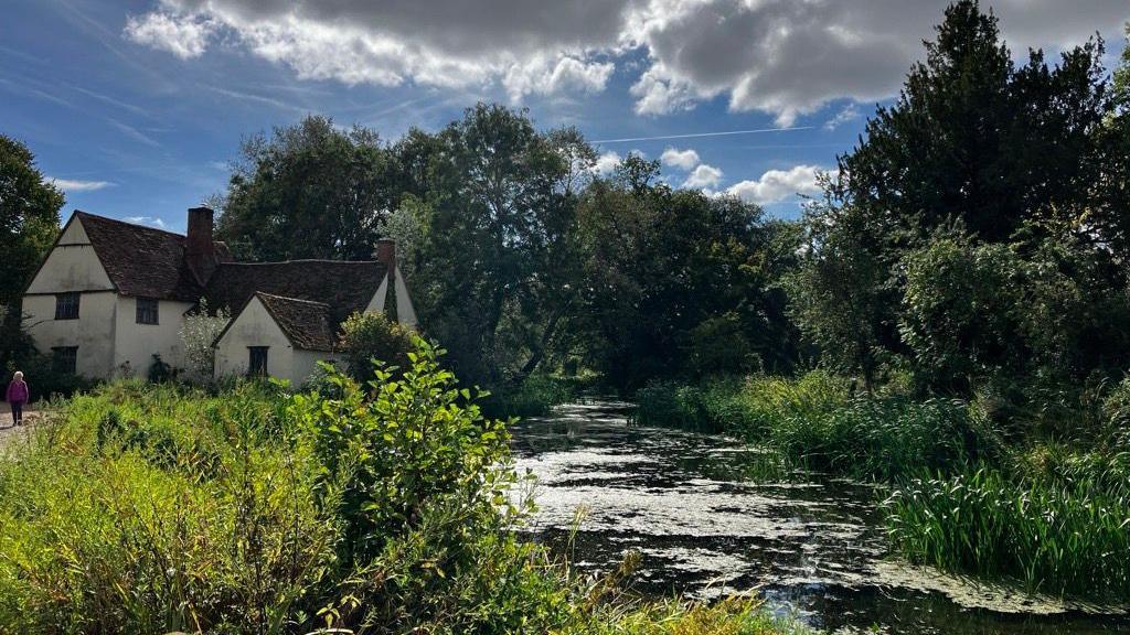 An idyllic scene in Flatford, near Dedham, featuring trees and showing the River Stour and a white cottage on the left river bank.
