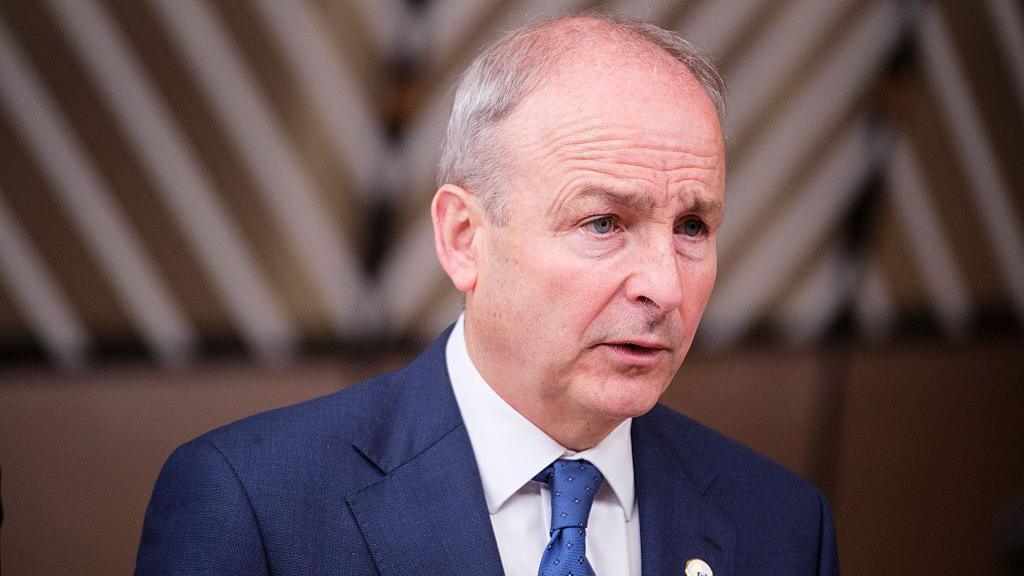 A balding grey-haired man wears a blue jacket with a white shirt and blue tie.