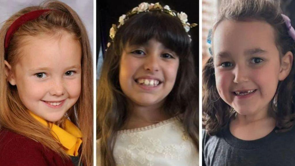 Headshots of three young girls. One on the left is dressed in a maroon school uniform and matching headband with fair long hair. The child in the middle is dressed in a white dress with a flowery headband and dark long hair. The child on the right wears a dark t-shirt with her hair tied in bunches. All three are smiling.