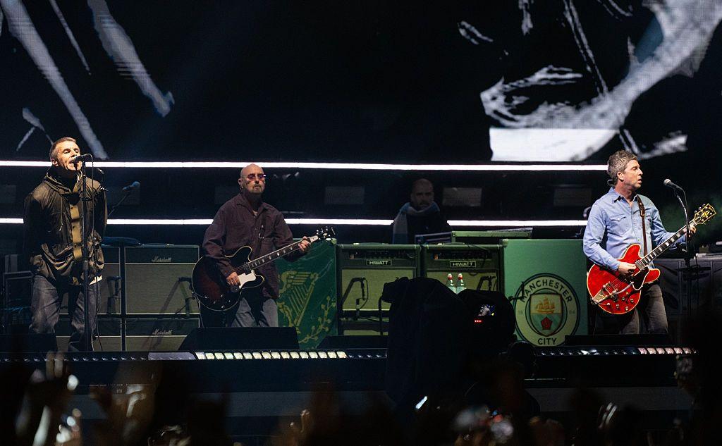 Liam and Noel Gallagher stand on stage at the Principality stadium. Noel (right) wears a denim shirt and holds a red guitar while Liam (left) wears a dark green parka. 