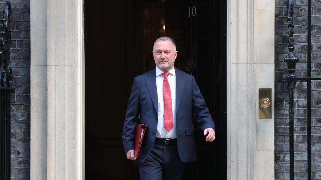 Steve Reed walks out of the black front door to No 10. He holds a red folder under one arm. He wears a navy suit, white shift and spotty red tie.
