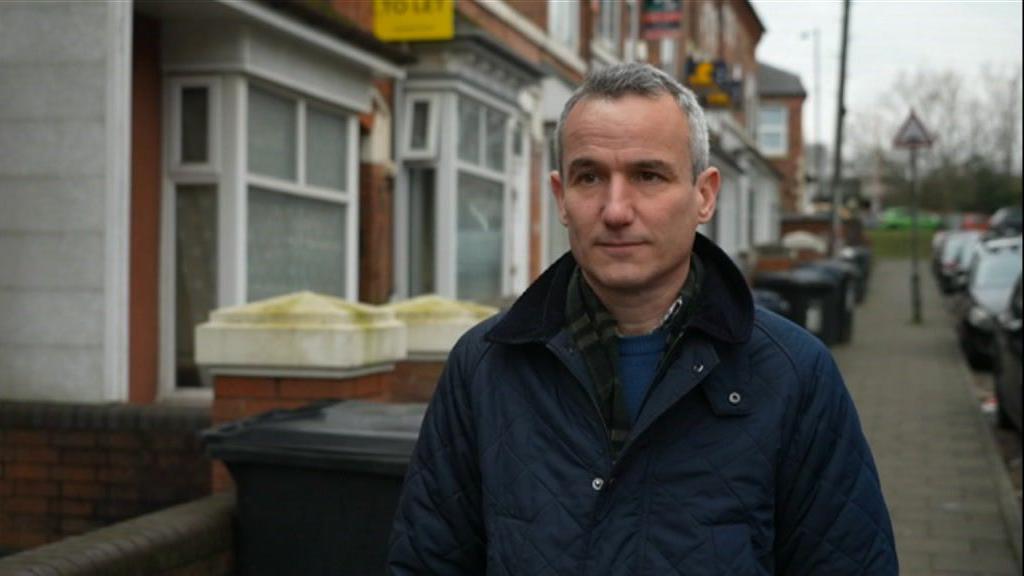 A man with short grey hair is wearing a dark blue jacket and is standing on a residential street with houses in the background.