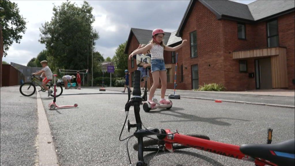 A little girl on a two wheel scooter with a pink helmet.