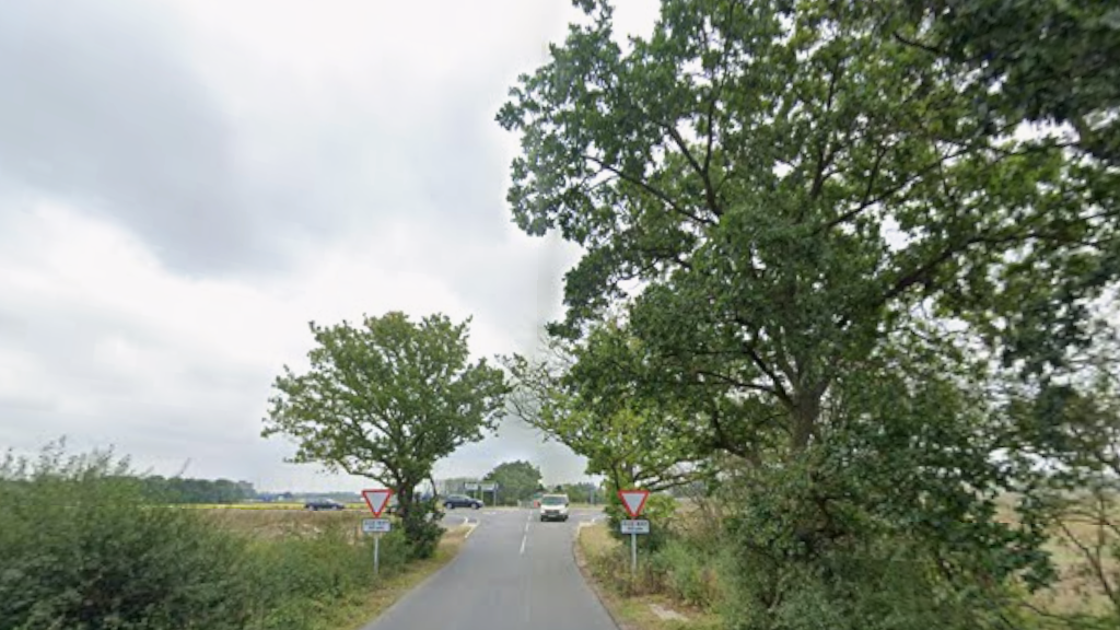 Part of a country road has trees and bushes on both sides. There are give way signs as a main road lies ahead. There is a van on the small road and two cars can be seen on the main road in the distance.
