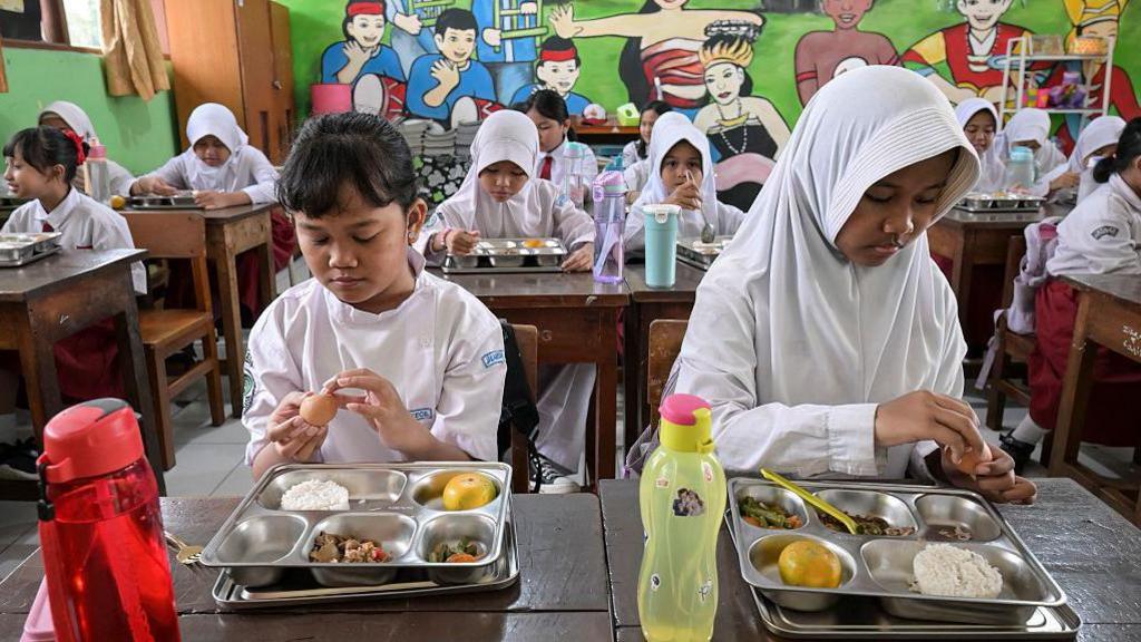 Two young girls eat from metal trays at a classroom in Indonesia, with their classmates doing the same in the background.