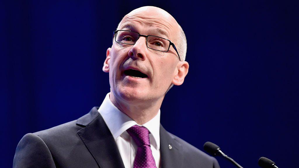 John Swinney, in a black suit and purple tie wearing glasses, is mid-speech. He stands in front of a microphone with a dark background.