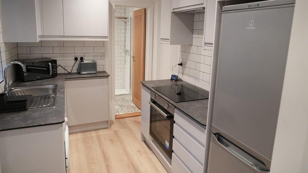 The kitchen inside a three-bedroom home in Carr Avenue, Leiston. It shows a light brown laminate floor. There is a grey fridge and a dark grey worktop. The kitchen units are an off white colour. 