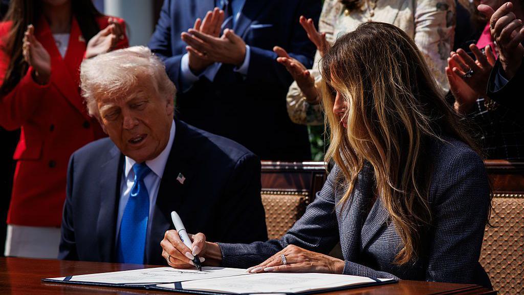 Donald and Melania Trump at a table in the Rose Garden of the White House, signing the "Take it Down Act" into law. She is wearing a dark grey suit and signing the document with a large black marker.