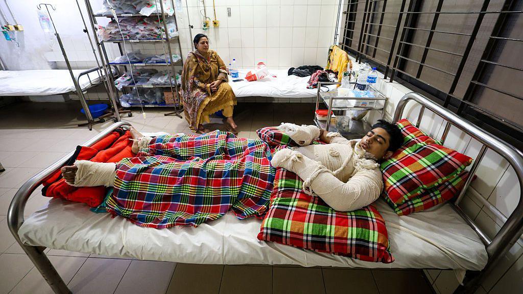 A boy covered in bandages, lying on a hospital bed. A woman in a yellow dress sits on the bed beside him and watches him.