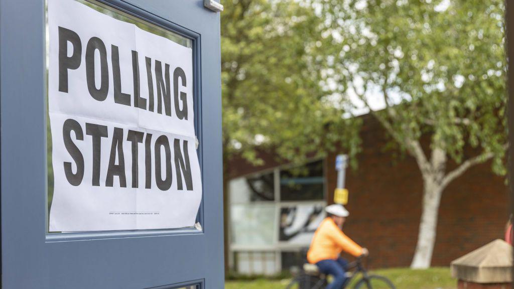 A polling station poster on the glass of an open door.- it reads "Polling Station" in black font on a white background. In the background there is a cyclist that is out of focus. They are wearing an orange coat and blue trousers and are cycling past two trees and a green lawn.