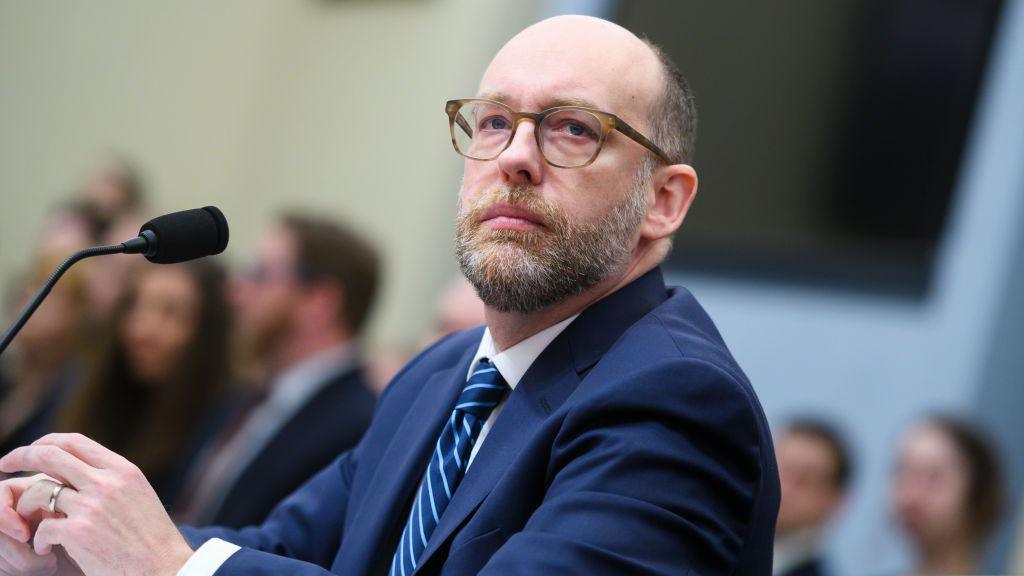 Russell Vought is seated during a hearing with a microphone direct toward him. He is wearing a blue suit and a stipped blue tie. He is wearing glasses. 