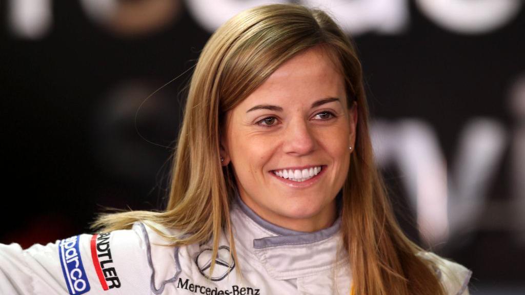 Susie Wolff in 2011 with long hair, smiling and wearing a white racing suit covered in sponsor badges. The back ground is out of focus.