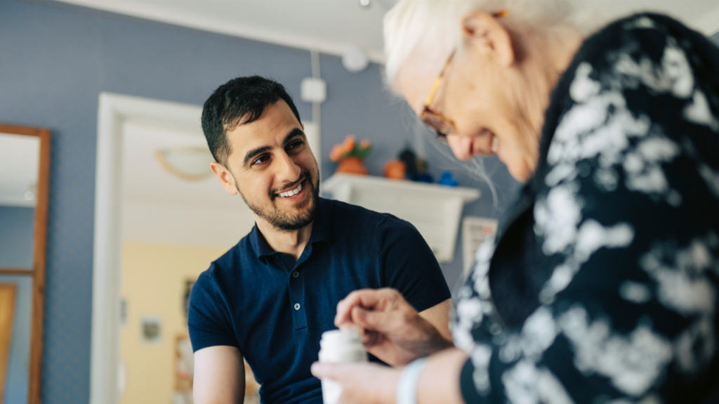 A smiling man in a blue polo shirt and trousers sits next to a smiling older woman who is taking pills out of a bottle