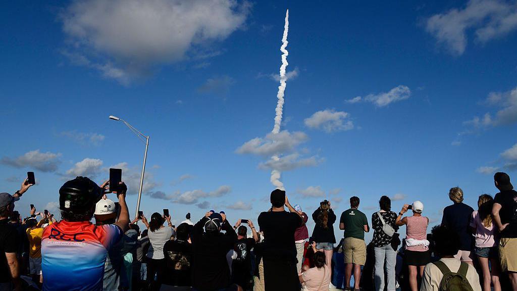 Crowd points phones to the sky as they take pictures of rocket take off