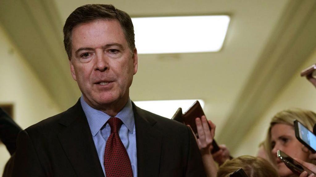 James Comey walking through a hallway where reporters are gathered 