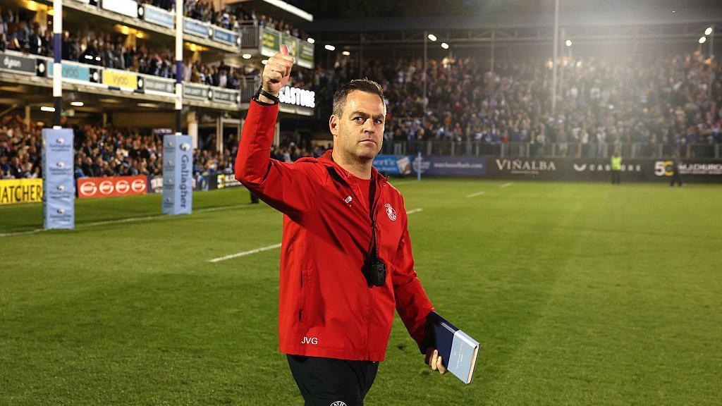 Johann van Graan holds his arm in the air and gives a thumbs up to the crowd while walking on the pitch at the Recreation Ground