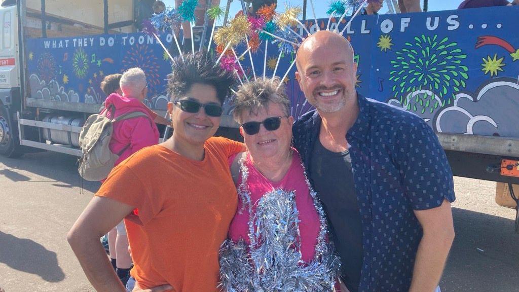 Three people stood in front of a float. One is wearing an orange shirt, one is wearing a sequined top and the other is wearing a blue top.