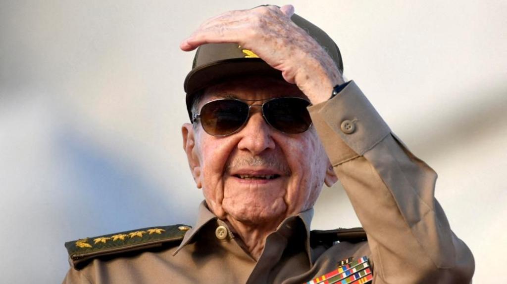 Raul Castro, wearing sunglasses and a military uniform, shields his face from the sun