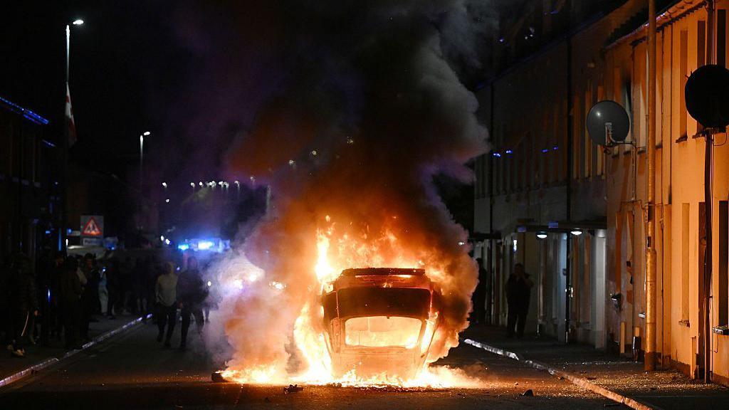 A car lying on its roof in the middle of a road. It is on fire. There are houses on either side of the street.