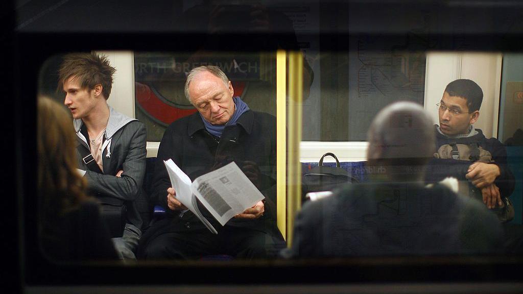 Ken Livingstone sits on a Tube train reading some documents. The photo was taken from outside the train looking in through the window. 