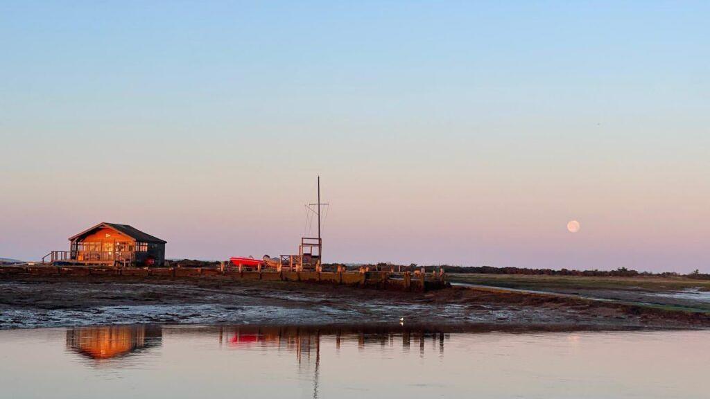 The banks of a river is illuminated by some wintery, low-light sunshine. A small cabin is visible on the bank and is bathed in orange light from the low sun. The cabin and a small jetty are reflected in the water and the moon is lightly visible in the top right hand corner.