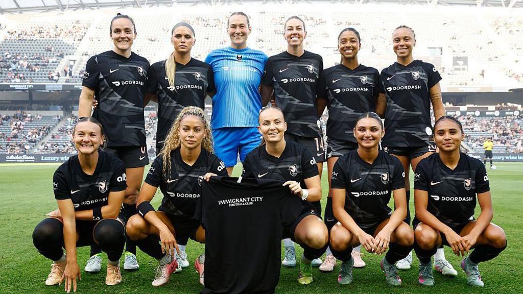 Angel City players pose for a photo with a shirt reading 'Immigrant City Football Club'