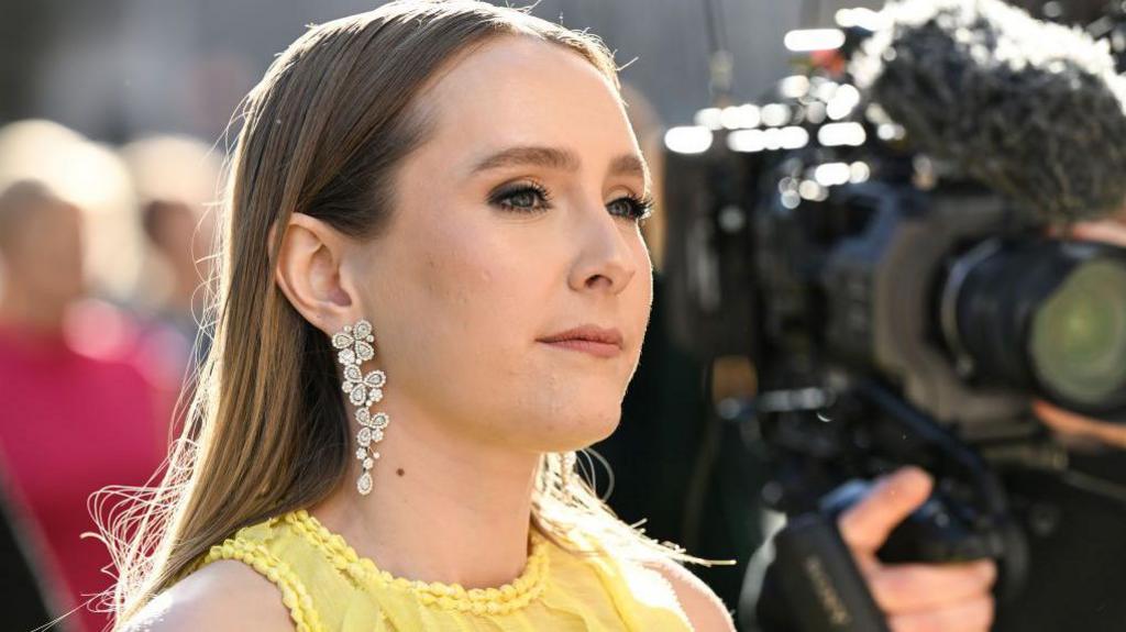 A headshot of Rose Ayling Ellis wearing a yellow dress and long sparkly earrings as she attends The Olivier Awards 2023 at the Royal Albert Hall. A camera and other blurred out figures can be seen behind her.