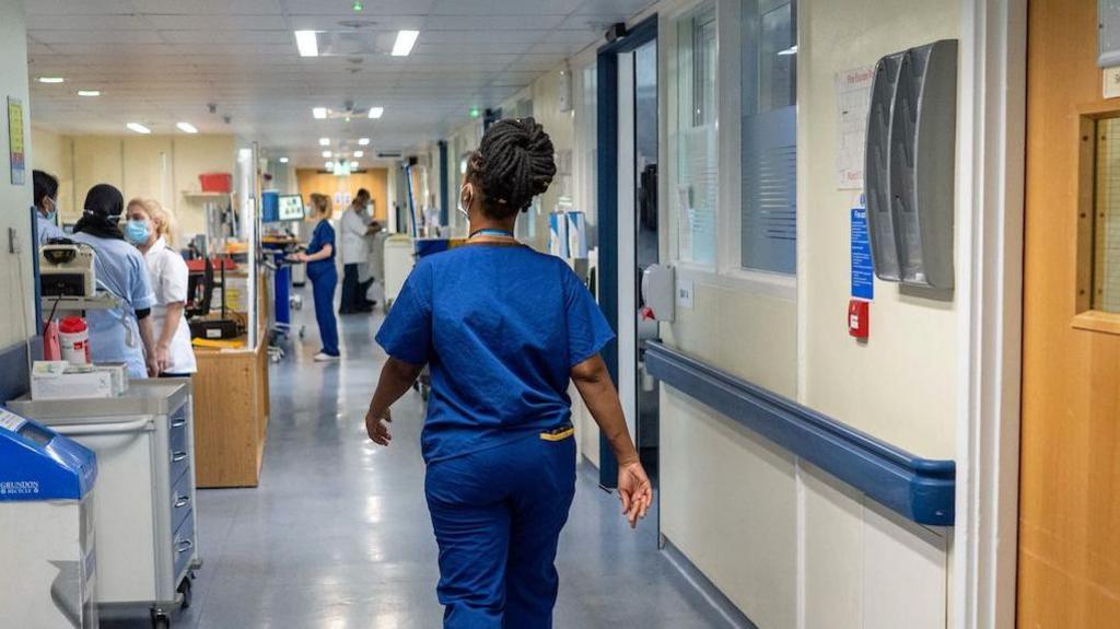 A nurse wearing blue scrubs walks down a hospital corridor. 