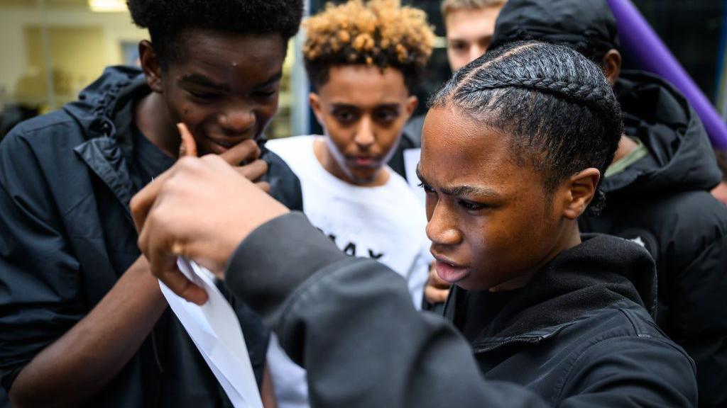 A student, who is wearing a black jacket and has a braid in his hair, looks quizzically at his GCSE results slip. His friends look on, one of them holding his hand to his mouth.