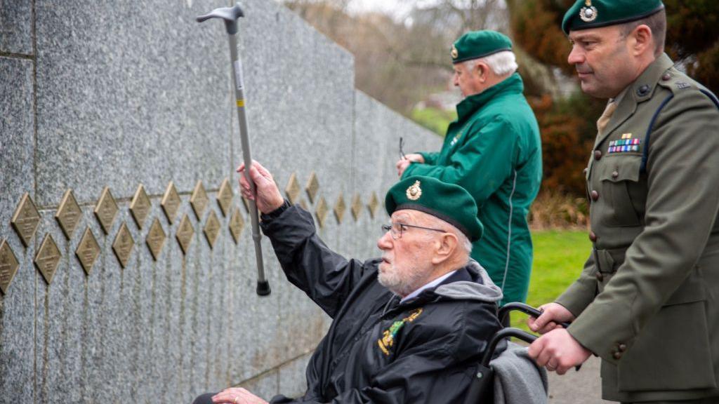 John Eskdale wearing a green beret while sitting in a wheelchair and holding up his walking stick as he looks at a memorial. He is accompanied by two other men. One in uniform and another wearing a green waterproof jacket.