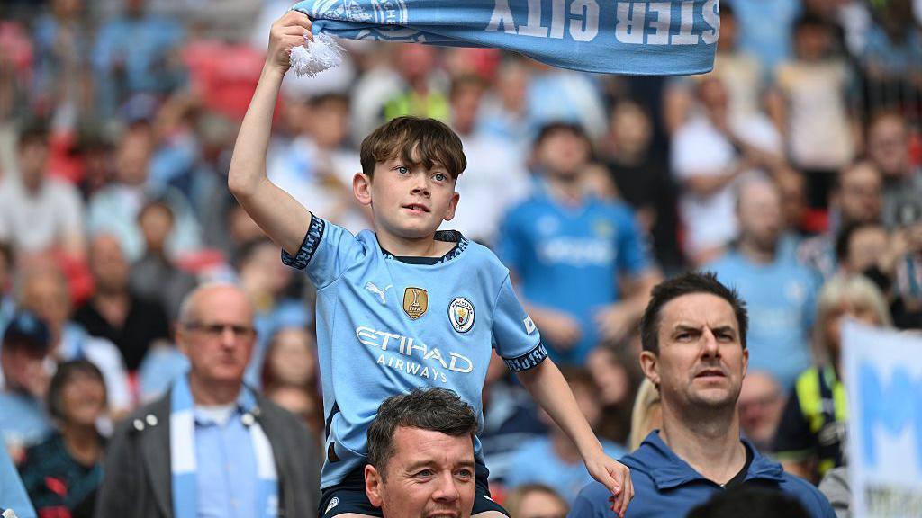 A young Man city fan waves a scarf above his head whilst on an older man's shoulders.
