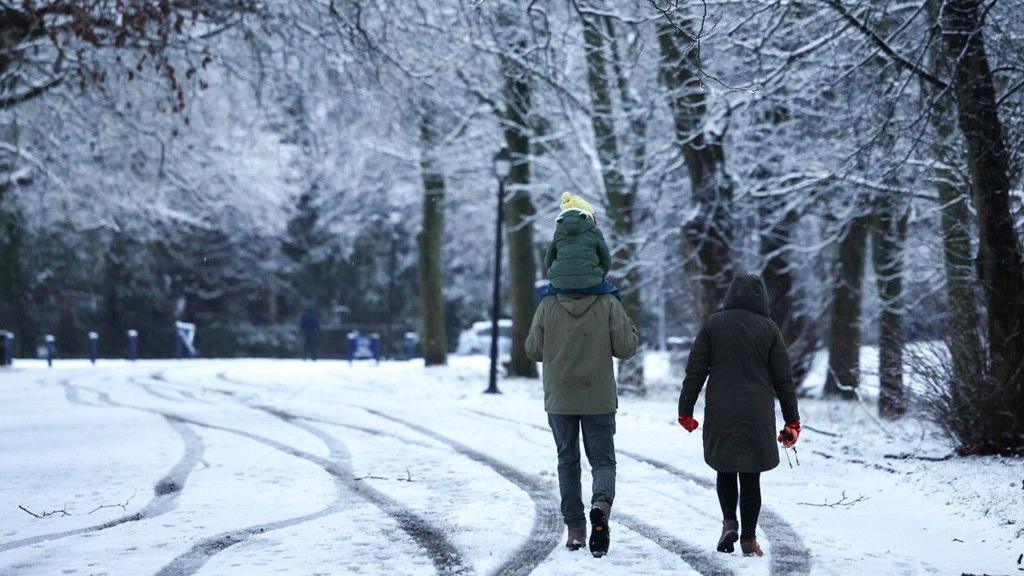 A couple walk down a snowy road. The man is carrying a child on his shoulders. There are big trees with some snow on them lining the road. 