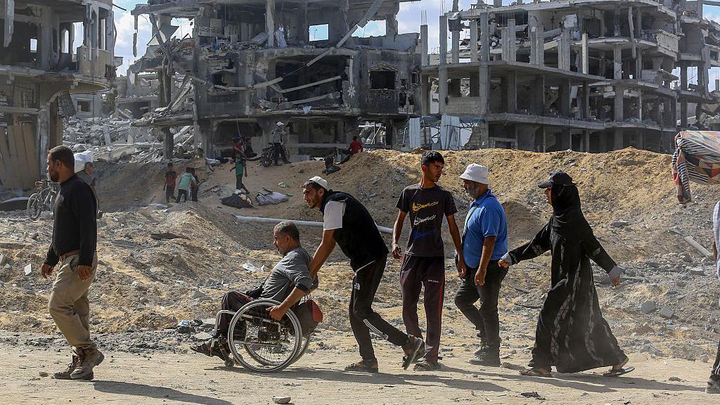 Palestinians walk in front of rubble in Gaza. One of the Palestinians is in a wheelchair, while someone behind him pushes him.