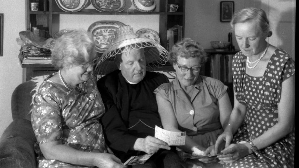 A black and white photograph of three women (Alice on the far right) sitting on a sofa reading postcards with a male vicar wearing a woven rattan hat. 