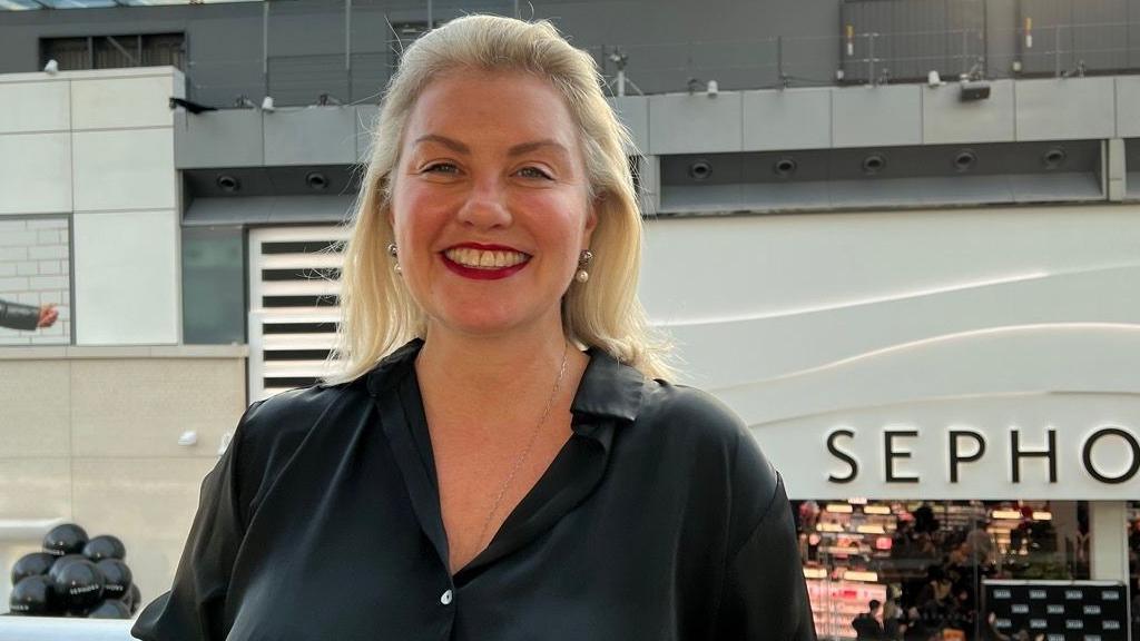 Sarah Boyd is wearing red lipstick, has blonde hair and is wearing a black shirt. She is standing in front of a shop entrance and the letters S E P H O are visible on a sign behind her