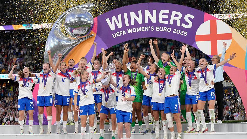 Lionesses celebrate in front of a banner that reads "Winners EUFA Women's Euro 2025" with gold confetti in the air. Their arms are raised and they are chanting.