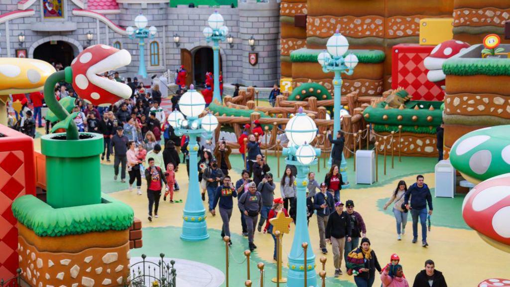 People walking in a theme park, with large mushroom plants and lamp posts either side of a walkway and a castle in the background