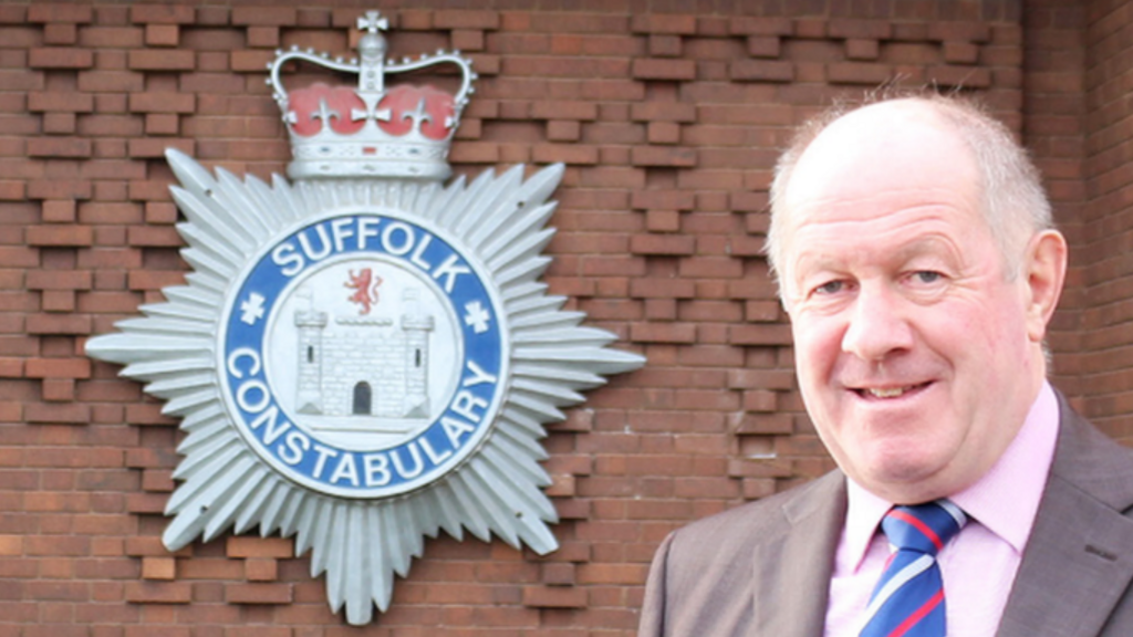 Tim Passmore, wearing a brown blazer, pink shirt and blue, red and white striped tie posing next to the Suffolk Constabulary badge