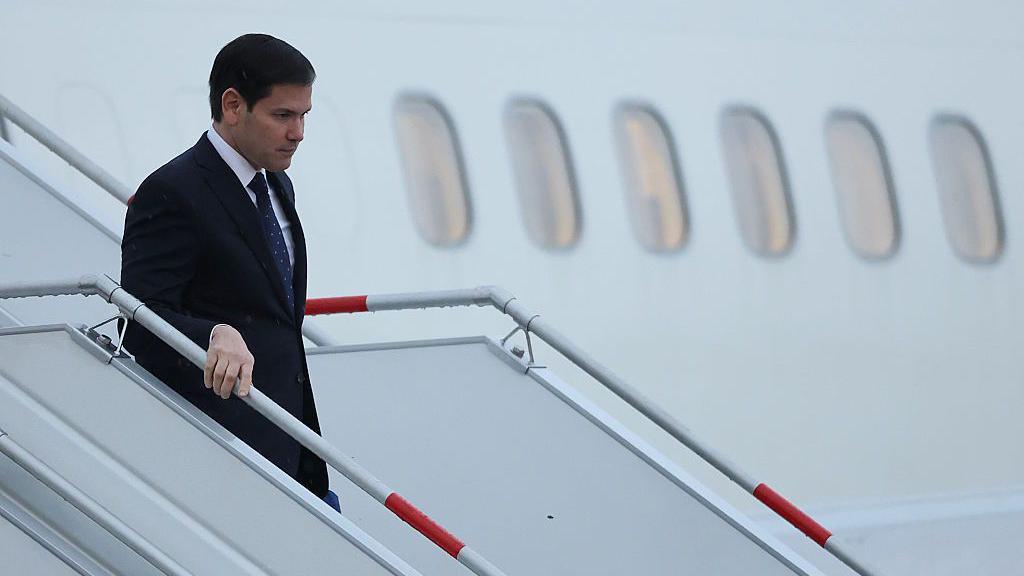 Marco Rubio walks off a plane holding a rail wearing a suit