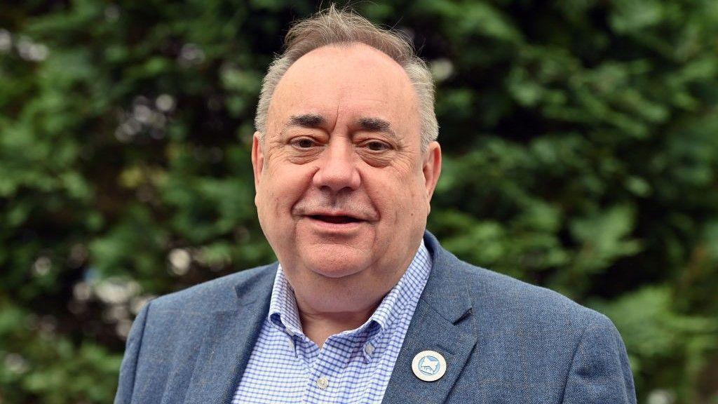 Alex Salmond smiling in medium close up. He is wearing a blue tweed jacket, open-necked button down white and blue checked shirt and an Alba party badge