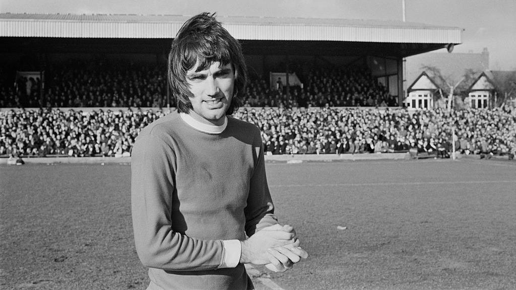 George Best facing the camera at a Manchester United match against Northampton in 1970. The black-and-white image shows him on the pitch with a full stand of supporters behind him. 