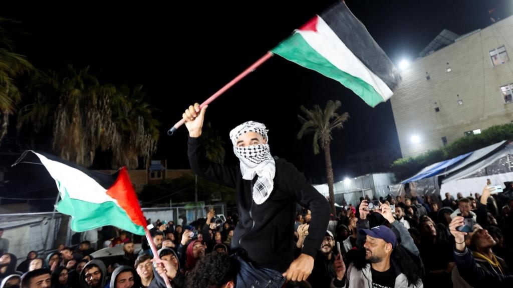 A man sits on another man's shoulders and waves a black, white, red and green Palestinian flags as people celebrate around him 