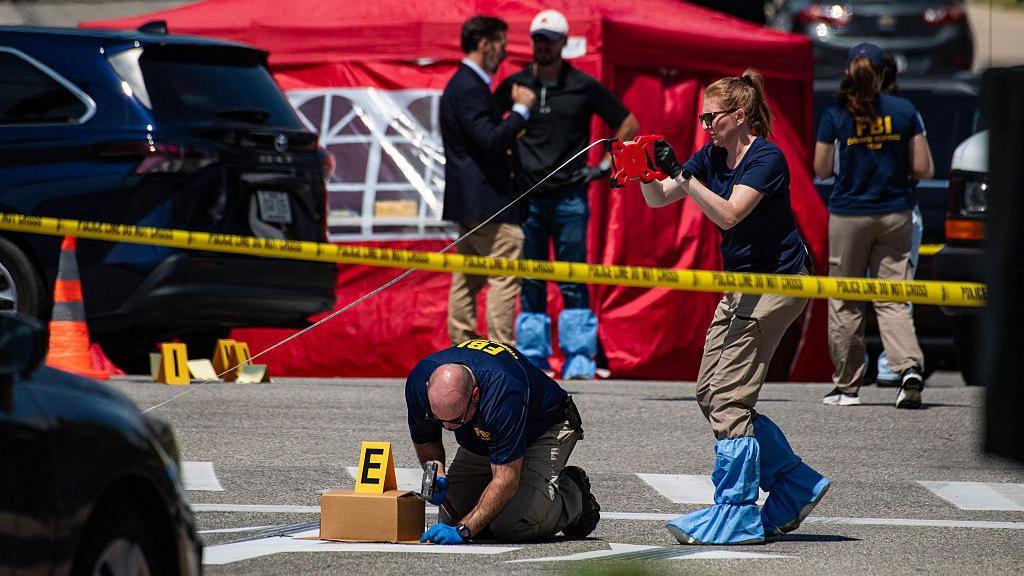 An image showing a pair of investigators behind caution tape surveying the scene of a shooting in Maine, where a 26-year-old man was shot and killed by an ICE agent. Behind them are an orange cone, a black van, other FBI investigators and a red tent.