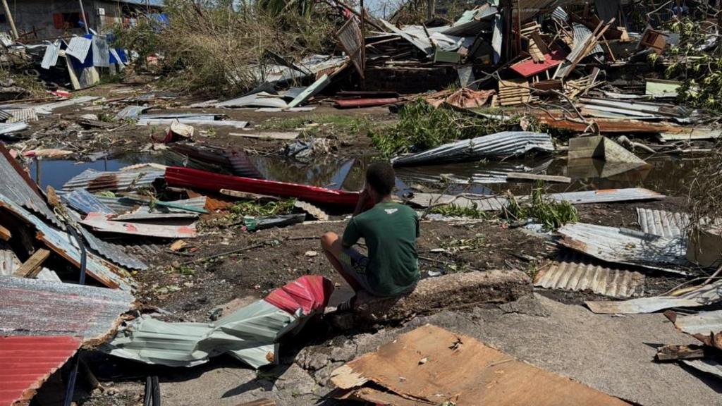 Young man in green t shirt sits facing away from the camera, with debris surrounding him as metal sheets and cardboard strewn all over the path and small stream of water near to him