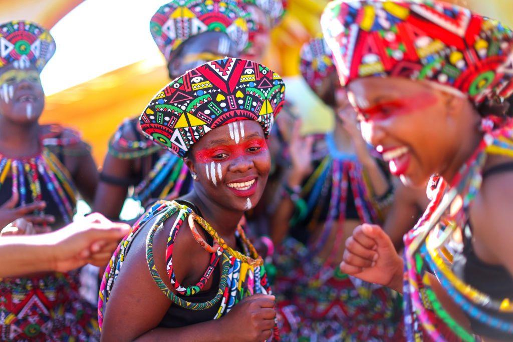 Carnival performers in traditional multi-coloured South African costumes and hats smile. They are also have a stripe of red face paint over their eyes and white markings painted on their cheeks, forehead and chin - Cape Town, South Africa - Wednesday 6 November 2024