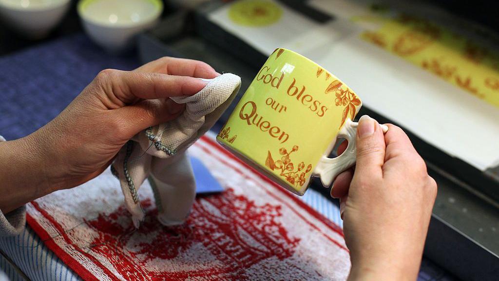 The hand of a ceramics worker seen polishing a yellow mug with red lettering saying God Bless Our Queen.