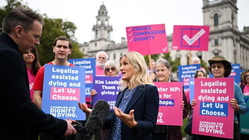 Kim Leadbeater, wearing a navy jacket and blue top, being interviewed by a journalist with a microphone. Behind her are a number of campaigners holding placards in support of the assisted dying bill. Their signs say "Legalise assisted dying and let us choose".