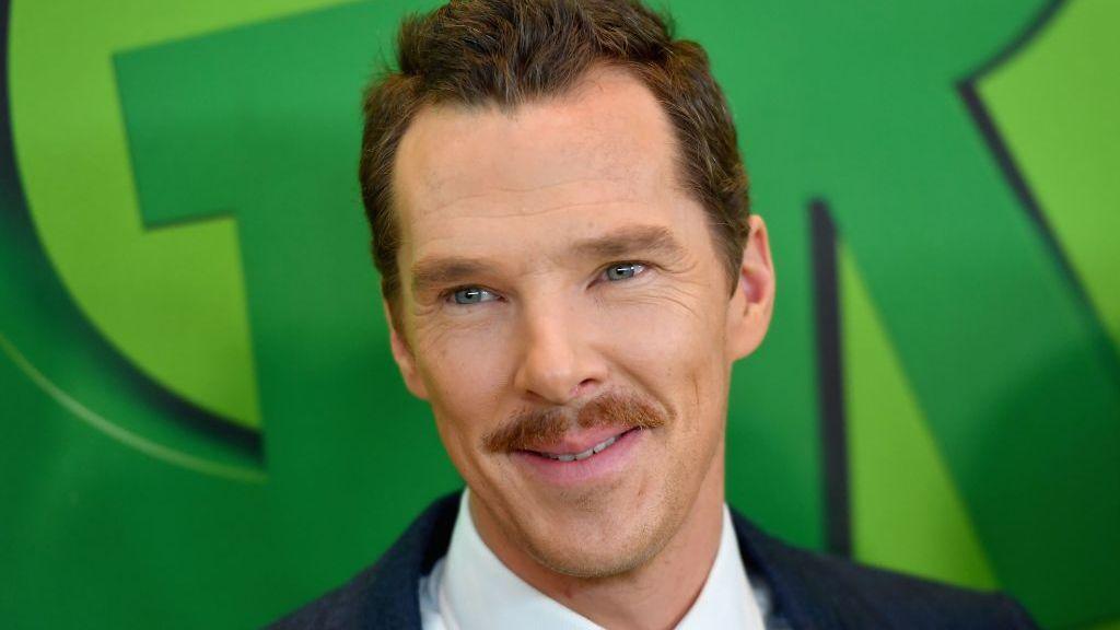 Benedict Cumberbatch looks away from the camera smiling. He has short brown hair and a brown moustache. He is wearing a dark suit and white shirt. he is standing in front of a green billboard that says 'The Grinch'.